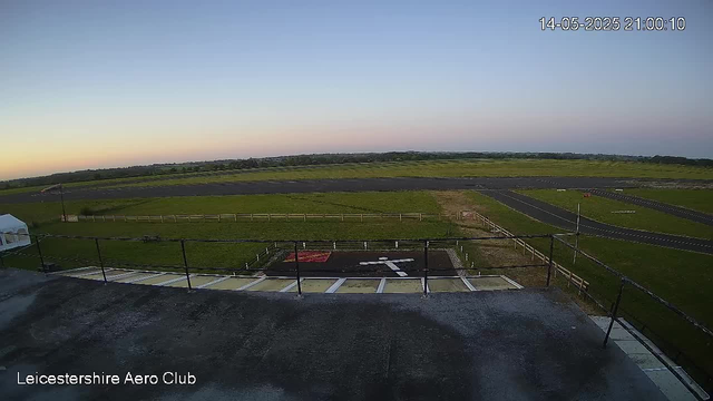 An outdoor view from a webcam showing a runway and grassy fields extending into the horizon under a clear sky during sunset or sunrise. There is a small helipad with a white arrow marking on it in the foreground, and a fenced area surrounding it. A flag is visible on the left side of the image, and the timestamp in the top right corner reads "14-05-2025 21:00:10."