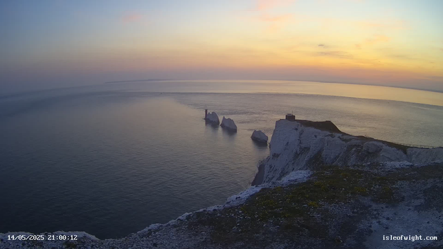A view of white chalk cliffs overlooking the ocean at sunset, with a lighthouse on the left side, a small building on the right, and a few scattered rocks in the water. The sky is a gradient of pastel colors, fading from pink to blue, with calm water reflecting the soft light.