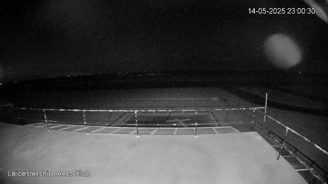 A black-and-white image from a webcam shows an empty outdoor sports field surrounded by a low fence. The scene appears to be nighttime, as the sky is dark. There is a digital timestamp in the top right corner showing the date and time as 14-05-2025 at 23:00:30. The bottom left corner has the text "Leicestershire Aero Club." There are some bright spots, potentially reflections or lens flares, visible in the image.