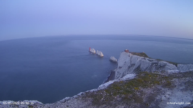 Aerial view of white chalk cliffs along a coastline, with a small lighthouse on one of the cliffs. Large white chalk stacks rise out of the blue ocean nearby. The sky is clear with a faint horizon line in the distance. The scene appears to be early morning.