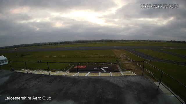 A wide view of a runway and grassy fields seen from an elevated perspective at Leicesterhire Aero Club. The sky is partly cloudy with some sunlight breaking through clouds. There is a small helipad marked with an "H" in the foreground, and the surrounding area is mostly flat open land. A timestamp in the top right corner shows "15-05-2025 05:00:51."