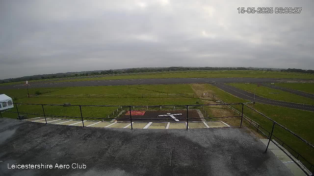 An overcast view of an airfield shows a runway and surrounding grassy areas. The image is taken from a tall vantage point, overlooking the tarmac and a section of parking spaces with a small helicopter-style cover. Faint clouds cover the cloudy sky, and the landscape extends into the distance with gentle hills on the horizon. The bottom-left corner displays the text "Leicestershire Aero Club." The timestamp in the top-right corner reads "15-05-2025 06:00:27."