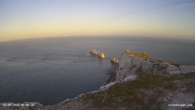 A view of white chalk cliffs over the ocean at sunrise, with two prominent rock stacks near the shore. The cliffs have a grassy top with some structures, and a lighthouse is visible on one of the rock stacks. The sea appears calm, and a ship can be seen far in the distance. The timestamp indicates the photo was taken early in the morning on May 15, 2025.