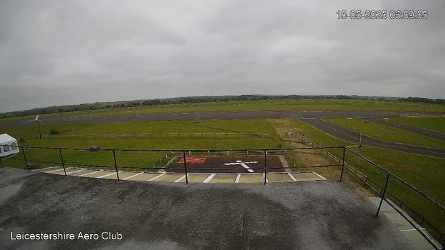 A view from a webcam showing an overcast sky with grey clouds. In the foreground, there is a concrete platform with a black railing, and a small area with painted markings and directional arrows. Below the platform, there is a parking area with designated spaces, one of which has an arrow painted on it. Beyond the parking area, there is a grassy field with a wooden fence and a flat, open landscape extending into the distance. The timestamp on the top right corner reads "15-05-2025 06:59:55." The bottom left corner has the text "Leicestershire Aero Club."