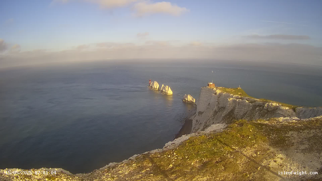 A panoramic view of the white chalk cliffs of Étretat, France, with tall, white rock formations extending into the sea under a partly cloudy sky. The foreground shows the edge of the cliff covered with green grass and moss. The ocean is calm, and a small building with a flag is visible atop the highest point of the cliffs. The timestamp at the bottom left indicates the image was captured on May 15, 2023, at 07:01:04.
