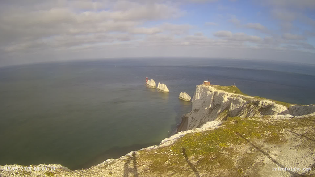 A panoramic view of white chalk cliffs with green grass on top, overlooking the ocean. There is a small structure with a red and white lighthouse on one of the cliffs. Several large rocky formations are visible in the water, and the sky is partly cloudy with patches of blue.