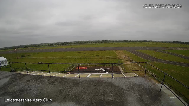 A view from a webcam showing the open sky with overcast clouds, a grassy area, and a runway in the distance. In the foreground, there is a balcony or platform with a metal railing, part of a building structure at the bottom of the image. The image is marked with the date and time "15-05-2025 08:00:54" in the top right corner and the text "Leicestershire Aero Club" at the bottom left corner.