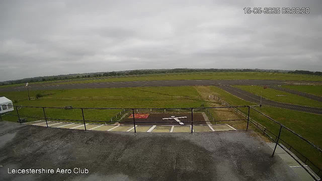 A wide view of an airfield showing a parking area with marked spaces and a small helipad, surrounded by grassy fields and a runway extending into the distance under an overcast sky. The scene is captured from a webcam at Leicestershire Aero Club, with a timestamp of 09:00:20 on May 15, 2025.