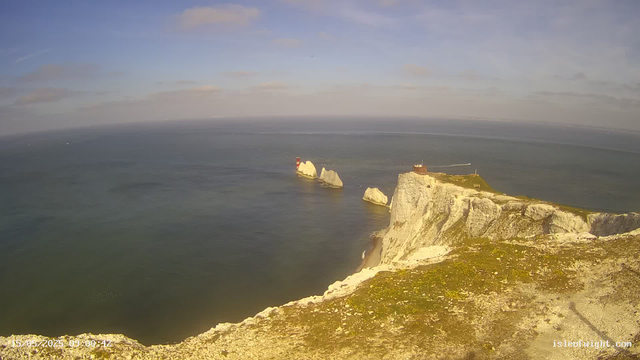 A panoramic view of white chalk cliffs along the coastline, with a small building and a lighthouse on top. The ocean stretches out below with a few large rocks emerging from the water, and a lighthouse with red and white stripes stands on a rock formation in the distance. The sky is mostly clear with some scattered clouds. The image was captured on May 15, 2023, at 09:00:42.