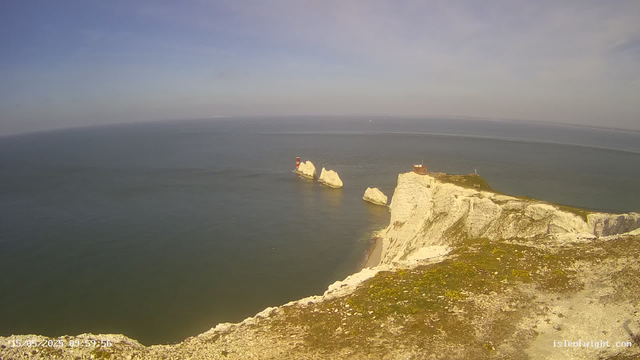 An aerial view of white chalk cliffs rising out of the ocean, with a lighthouse situated on one of the cliffs. Several large chalk rock formations are visible in the water near the cliffs. The sky is partly cloudy with a soft, diffuse light. The scene appears calm with minimal waves.