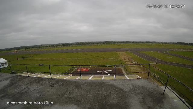 Overcast sky overlooking an airfield with taxiways and runways stretching into the distance. In the foreground, there is a parking area with marked spaces, a small patch of landscaped ground, and a railing. The timestamp in the upper right corner shows the date and time as May 15, 2025, 10:00:51. Text at the bottom left reads "Leicestershire Aero Club."