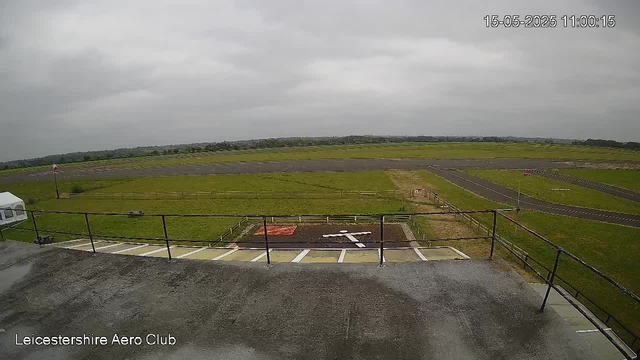 A view from a webcam showing an overcast sky above a grassy airfield with runways and taxiways in the distance. There is a parking area with some marked spaces and a small helicopter landing pad in the foreground. A wooden fence surrounds part of the grassy area, and the scene is labeled "Leicestershire Aero Club" in the bottom left corner. The timestamp in the top right corner reads "15-05-2025 11:00:15".