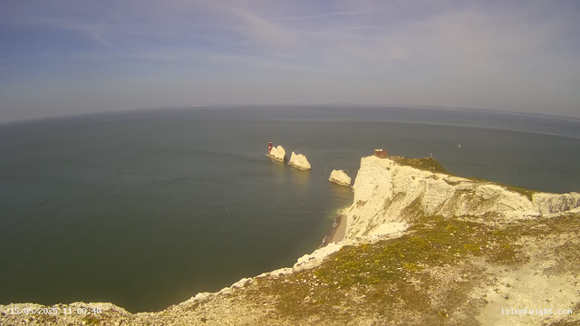 A panoramic view of white chalk cliffs along the coastline with a lighthouse standing on one of the cliffs. The sea extends to the horizon, with a few small boats visible in the water. The sky is mostly clear with some wispy clouds. The photo appears to have been taken in the daytime, and there's a timestamp at the bottom left corner.
