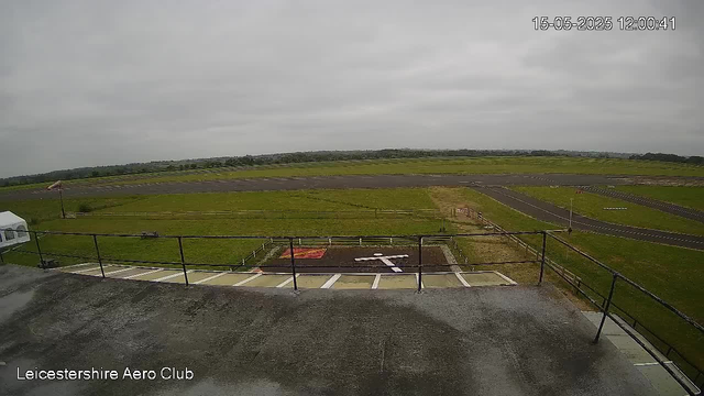 An outdoor view from a webcam at Leicesterhire Aero Club, showing a cloudy sky, a grassy airfield with a small runway, and a parking area with several parking spaces. The timestamp indicates the date and time as May 15, 2025, at 12:00:41.
