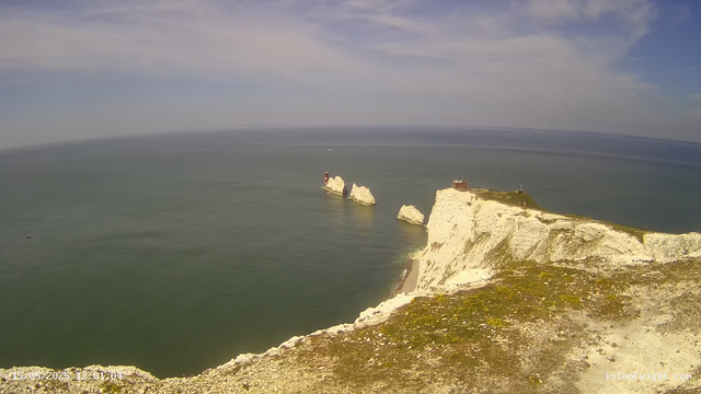 A view of white chalk cliffs extending into the ocean with large rock formations and a lighthouse on one of the cliffs, under a partly cloudy sky.