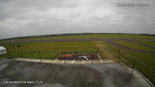 This image shows a view from a webcam at Leicestershire Aero Club. In the foreground, there is a rooftop area with a railing and some parking spaces. Beyond that, there is a grassy area and a small runway or taxiway with a white arrow painted on it. The scene is overcast with gray clouds, and the landscape stretches out into the distance with fields and low hills under a cloudy sky. The date and time displayed are 15-05-2025 at 12:59:55.
