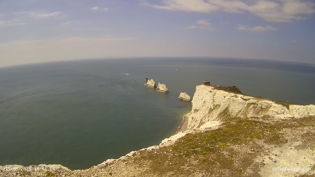 A panoramic view from a webcam shows white chalk cliffs extending into the sea, with a small building and grassy area on top of the cliffs. Several large rock formations rise from the water near the cliffs. The sea is calm with sailboats and a boat visible in the distance. The sky is partly cloudy with patches of blue. The timestamp at the bottom left reads "15-05-2023 14:01:04".