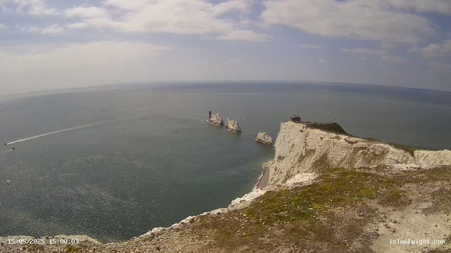 A high-angle view of white chalk cliffs with a grassy top, overlooking the ocean. In the distance, there are distinctive sea stacks with a lighthouse on the tallest one. A small red building is situated on the cliff's edge. The sky is partly cloudy, with patches of blue visible. A boat is moving across the water, leaving a wake behind it. The timestamp in the bottom left corner indicates the date and time as May 15, 2025, at 15:00.