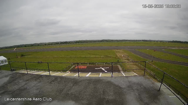 An outdoor view from a webcam showing an open field with a small taxiway or runway, a grassy area, and some fencing. The sky is overcast with gray clouds. In the bottom left corner, there is a logo that reads "Leicestershire Aero Club." The timestamp on the image is "15-05-2025 15:00:20."