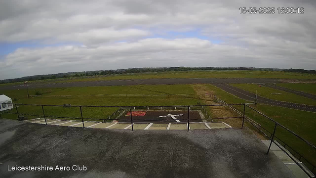 A view from a webcam at Leicestershire Aero Club shows an open grassy field with a runway stretching into the distance. The sky is partly cloudy with patches of blue, and the time and date are displayed in the upper right corner as 15-05-2025 16:00:12. The foreground includes a balcony or observation deck with a metal railing, and the bottom left corner has the text "Leicestershire Aero Club".