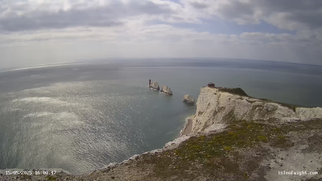 An aerial view of white chalk cliffs rising from the ocean, with a small building or lighthouse at the edge of the cliff. The sea below sparkles under a partly cloudy sky, and some rock formations are visible in the water near the base of the cliffs. The scene is peaceful and expansive.