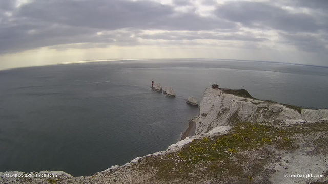 A wide view of white chalk cliffs along a coastline under a cloudy sky. Several large rock formations are visible in the water near the cliffs, with one lighthouse standing on a rock and a small building on top of the cliff. The date and time stamp at the bottom left corner indicates "15/05/2025 17:00:16," and the website "isleofwight.com" is displayed at the bottom right corner.