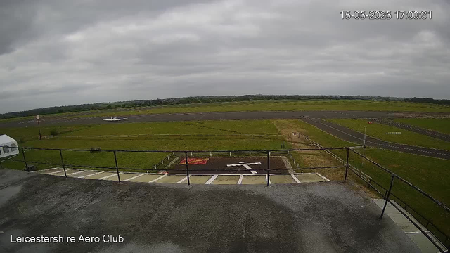 A view from a webcam showing an outdoor area of Leicester Shire Aero Club. In the foreground, there is a balcony with a black metal railing and a parking area with marked spaces. Beyond the balcony, there is a grassy field with a small aircraft parked near the runway. The runway extends across the middle of the image, with a paved taxiway running parallel. The sky is overcast with gray clouds, and there are no people visible in the scene. The top right corner displays a timestamp, and the bottom left corner has the text "Leicestershire Aero Club."