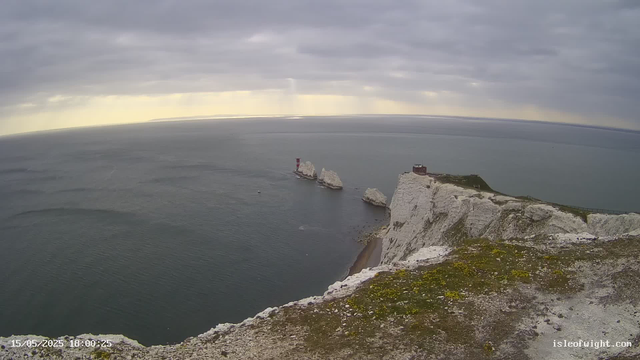 A view from a webcam showing white chalk cliffs extending into the ocean with a small building on top. There are three isolated rock formations in the water near the cliffs. The sky is overcast with thick gray clouds, and the sea appears calm. The image has a timestamp in the lower left corner showing "15/05/2025 18:00:25" and a website watermark in the lower right corner that reads "isleofwight.com."
