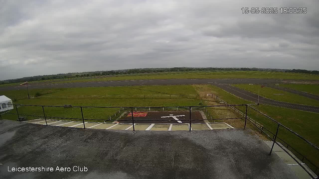 An overcast sky casts a gray hue over an open airfield with a large grassy area and a runway stretching into the distance. In the foreground, a section of a rooftop with a black railing is visible, along with a helipad marked with a white helicopter symbol. The image has a timestamp in the top right corner showing the date and time as 15-05-2025 18:00:35, and the bottom left corner indicates "Leicestershire Aero Club."