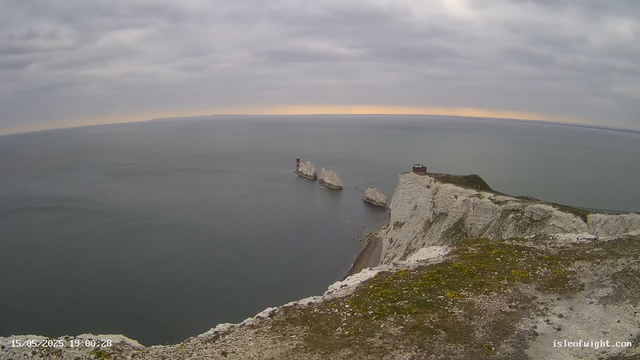 A view from a webcam shows white chalk cliffs extending into the ocean with several large white chalk stacks rising from the water. There is a lighthouse on one of the cliffs near the stacks and a small building nearby. The sky is overcast with gray clouds, and the sea appears calm. A timestamp in the lower left corner indicates the date and time as May 15, 2025, at 19:00:28. The website "isleofwight.com" is displayed in the lower right corner.
