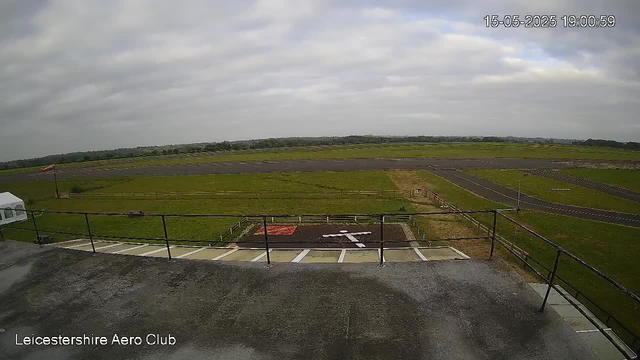 A wide view of a grassy airfield seen from a balcony or rooftop, with a parking area in the foreground and a taxiway leading to the runway. The sky is mostly cloudy with some patches of blue. There is an American flag on a flagpole on the left side. The image is timestamped "15-05-2025 19:00:59" in the upper right corner, and "Leicestershire Aero Club" is displayed in the lower left corner.