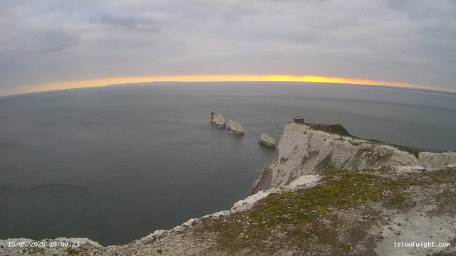 An aerial view of white chalk cliffs overlooking the ocean at sunset. Several large chalk rock formations are visible in the water near the shore, and a small building is situated atop the cliffs. The sky is mostly cloudy with a hint of orange and yellow near the horizon. A path runs along the edge of the cliffs. The timestamp on the image indicates it was taken on May 15, 2025, at 20:00.