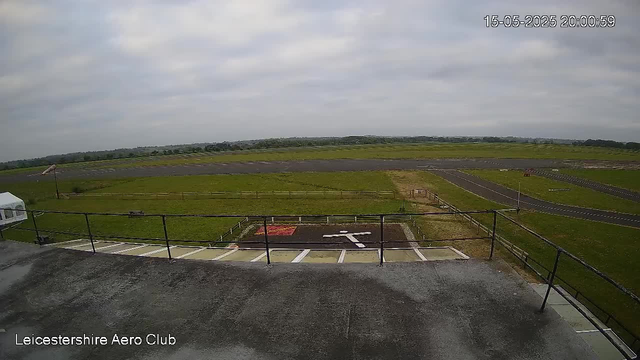 A view of an airfield with a grassy area and runways stretching into the distance. The image is taken from an elevated position, possibly a control tower. There are parking spaces and a helicopter landing pad in the foreground. The sky is cloudy, and there are few distant trees and structures on the horizon. The timestamp on the image shows the date and time as 15-05-2025 at 20:00:59. The bottom left corner has text that reads "Leicestershire Aero Club."