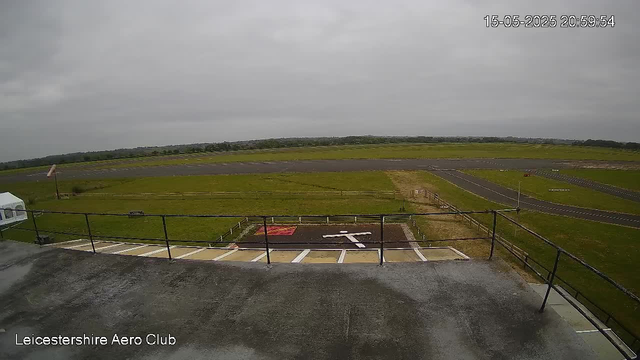 A view from a webcam at Leicester Shire Aero Club showing a cloudy sky over an open grassy area with a paved runway in the background. The image features a terrace or balcony with a metal railing in the foreground and some parking spaces below. The date and time stamp in the upper right corner reads "15-05-2025 20:59:54."