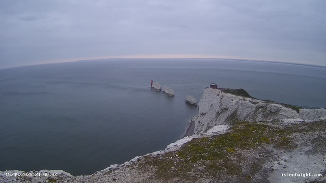 A view of white chalk cliffs extending into the sea, with isolated rock formations in the water. A small red and white lighthouse stands on one of the rocks, and a building is perched on the cliff top. The sky is overcast with gray clouds, and the ocean appears calm. There is a pathway along the edge of the cliff on the right side of the image.