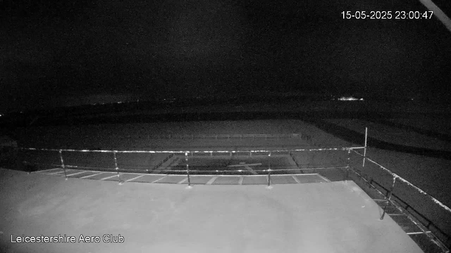 A black-and-white image taken at night from a webcam at Leicester-shire Aero Club. The foreground shows a flat, open area with a railing around what appears to be a small aircraft or platform. The background is dark with no visible details, and the sky is completely black. The timestamp in the upper right corner reads "15-05-2025 23:00:47."