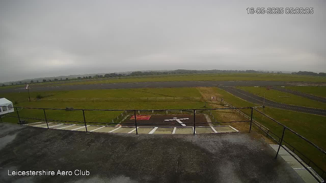 The image shows an outdoor scene at Leicesterhire Aero Club, viewed from a webcam. In the foreground, there is a helipad with a helicopter landing mark and surrounding safety markings, protected by a black metal railing. To the left, a small white tent is partially visible. Beyond the helipad, there are grassy fields, a few narrow pathways, and a small airstrip with runway markings extending into the distance. The sky is overcast with thick gray clouds, suggesting cloudy weather. The timestamp in the upper right corner indicates the photo was taken on May 16, 2025, at 5:00 AM.