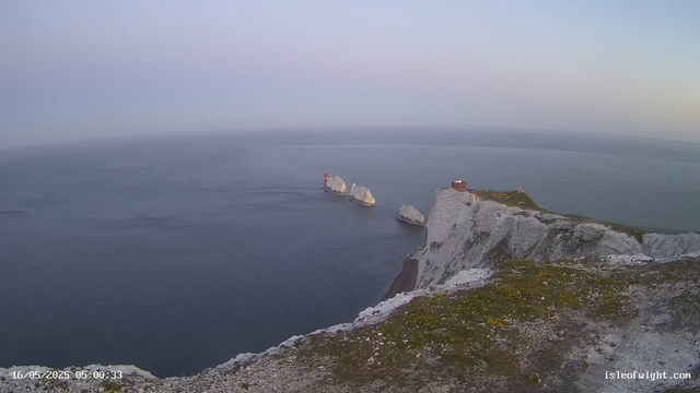 A view from a high vantage point overlooking the white chalk cliffs and the sea below. There are several large rock formations in the water, with one having a red and white lighthouse on top. On top of the cliffs, there is a small building surrounded by patches of grass and a walking path. The sky is clear, and the scene appears to be at sunrise or sunset. The timestamp shows the date and time as May 16, 2025, at 5:00 AM.