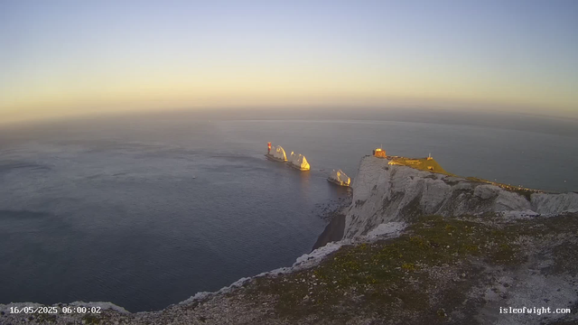 An aerial view of white chalk cliffs overlooking the ocean at sunrise or sunset. Several small icebergs or rock formations are visible in the water near the cliffs, with a lighthouse or tower on a rock in the distance. The sky is clear with a gradient of light, and the water appears calm. The date and time stamp in the bottom left corner reads "16/05/2025 06:00:02," and the website "isleofwight.com" is visible in the bottom right corner.