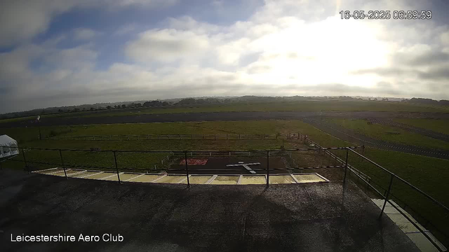 A wide view of an airfield or airport runway taken from a webcam, showing a grassy landscape, a runway in the distance, and a partly cloudy sky with the sun shining brightly. The image has a timestamp of 16-05-2025 06:59:59 and the text "Leicestershire Aero Club" in the bottom left corner.