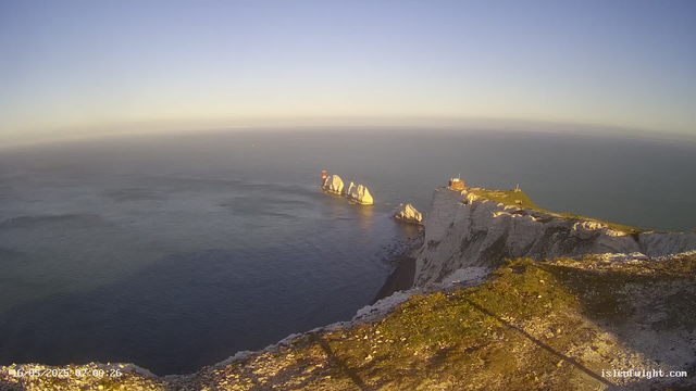 A scenic view of white chalk cliffs with patches of green grass on top, overlooking a calm sea. In the distance, two rocky islands with a lighthouse on one of them can be seen. The sky is clear with a gradient of blue, and the sunlight illuminates the landscape, suggesting it is either early morning or late afternoon.
