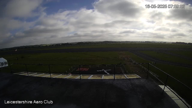 A wide view of a grassy airfield with a runway stretching into the distance under a partly cloudy sky. There is a small fenced area on the ground, and a transparent structure can be seen on the left side. The timestamp on the top right indicates the date and time as May 16, 2025, at 07:59:52. The text "Leicestershire Aero Club" appears at the bottom left of the image.