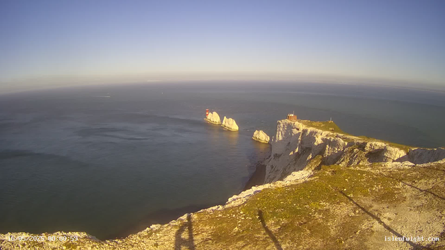 A view from a webcam showing white chalk cliffs along the coast with a grassy top, a small building, and a lighthouse on the right side. Several large rock formations are visible in the water below, and the sea extends to the horizon under a clear sky. The shadows of the person taking the photo are cast onto the ground in the foreground.