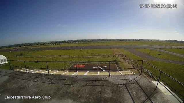 A wide-angle view of an airfield with a grassy runway area, taxiways, and a small set of stairs with a railing in the foreground. The sky is clear with a few small clouds, and the sun is shining from the top right corner. There is a flag on a pole near the edge of the grassy area and some fencing around the perimeter. The image includes a timestamp in the top right corner reading "16-05-2025 09:00:48" and the bottom left corner has the text "Leicestershire Aero Club."