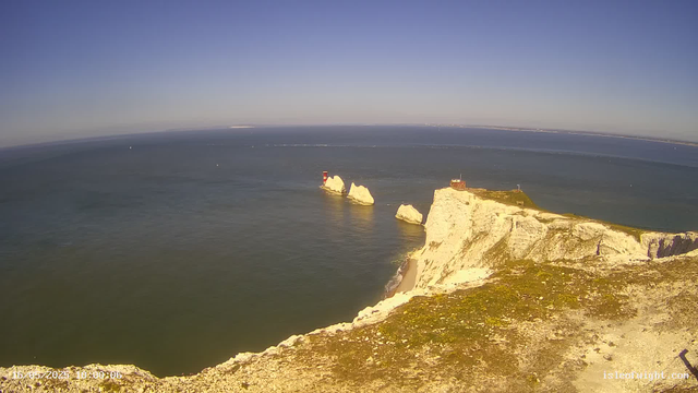 A high-angle view of a coastal landscape featuring white chalk cliffs with a grassy top extending into the ocean. Several large chalk stacks rise from the water near the base of the cliffs, with one marked by a small red and white lighthouse. A small building and what appears to be a lighthouse or observation point are situated on the highest point of the cliff. The sea is calm, and the sky is clear with a deep blue hue. The image appears to be captured from a webcam high above the coast during daytime.