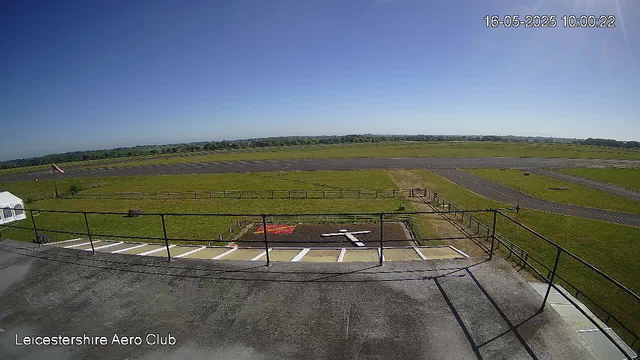 A wide view of an airfield shows a large open grassy area with taxiways and runways in the distance. The sky is clear and blue. In the foreground, there is a parking area with white lines, a small tent on the left, and a red helicopter markings on the ground near the fence. The timestamp in the top right corner reads "16-05-2025 10:00:22," and the text at the bottom left says "Leicestershire Aero Club."