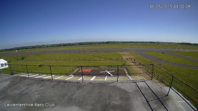 A wide-angle view from a webcam shows an open grassy airfield under a clear blue sky. In the foreground, there is a small parking area with white lines and a red-painted section, along with a metal railing. The bottom left corner displays the text "Leicestershire Aero Club." In the background, the runway extends into the distance, surrounded by green fields. The image is timestamped "16-05-2025 11:00:08" in the top right corner.