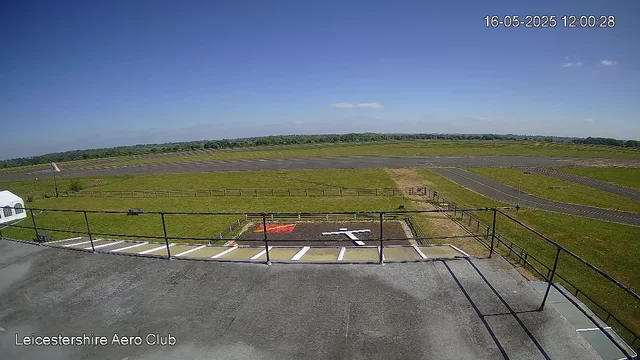 An outdoor view from a webcam at Leicestershire Aero Club shows an open grassy area with a pathway and a small parking lot. In the background, there is a runway extending across the scene, with a bright blue sky and a few scattered clouds overhead. The timestamp indicates the date as May 16, 2025, at 12:00:28.