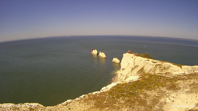 A view from a webcam showing white chalk cliffs overlooking the sea, with the iconic formation known as the Old Harry Rocks. A lighthouse is visible on one of the rocks, and there are some sailboats in the distance under a clear, blue sky.