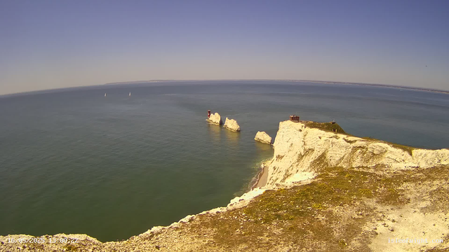 A wide view of white chalk cliffs extending into the sea, with a small building on top of the highest point of the cliffs. The ocean is calm with a few sailboats visible in the distance under a clear blue sky.